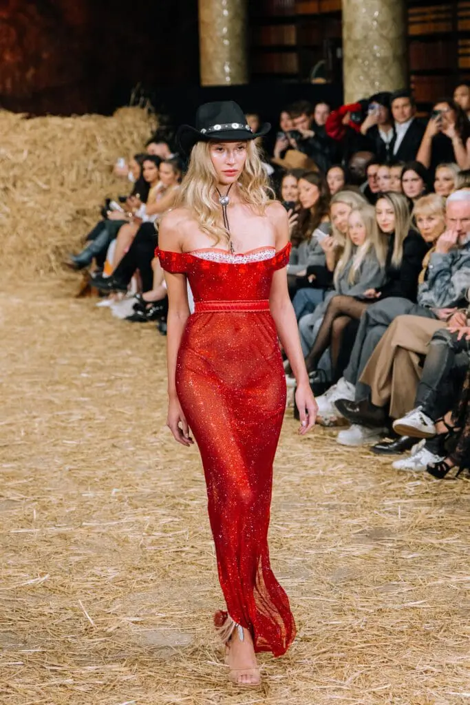 Model walking in red off-shoulder crystal gown with black cowboy hat at AADNEVIK AW25