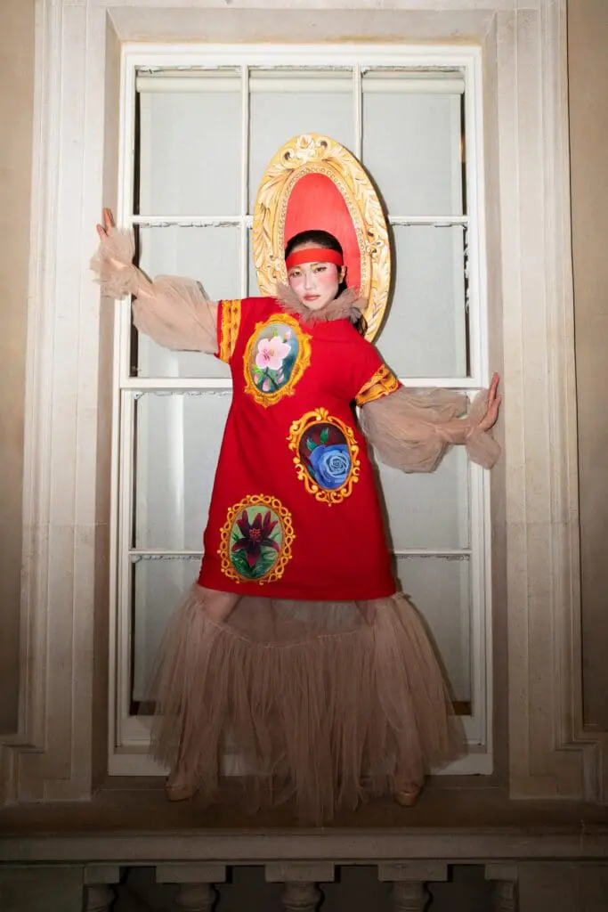 Model framed by an ornate gold frame in a red dress with floral appliqués and nude tulle