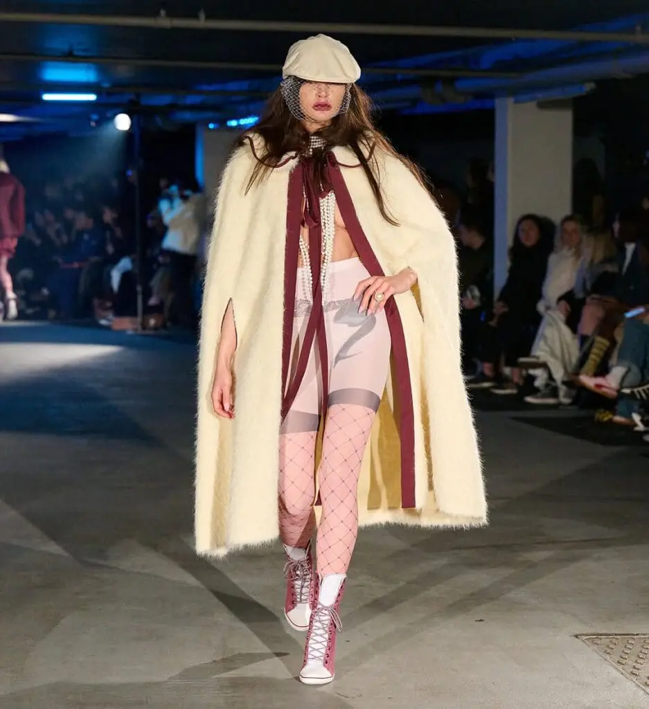 Model in oversized cream cape, sportswear-inspired shorts, and pink fishnet tights at Sinead Gorey AW25