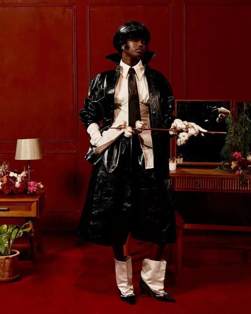 Model in a black glossy trench and tie, holding cotton branch like a staff