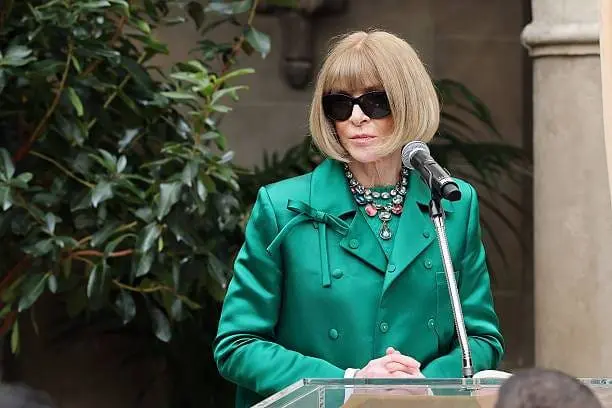 Anna Wintour in green coat and sunglasses speaking at podium