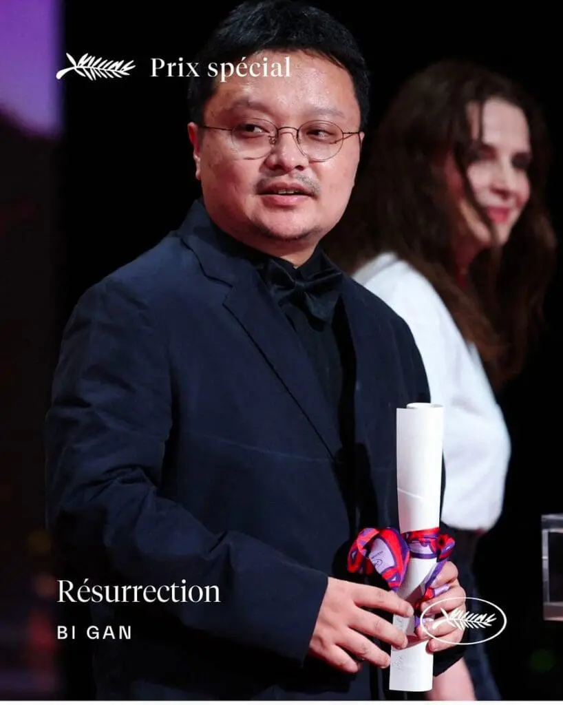 Bi Gan holding award scroll onstage with Palme d’Or backdrop