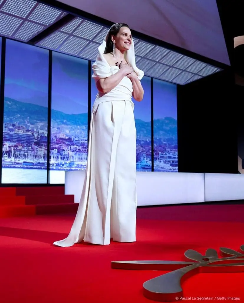 Juliette Binoche on stage at Cannes 2025 wearing a white gown and smiling with hands over heart