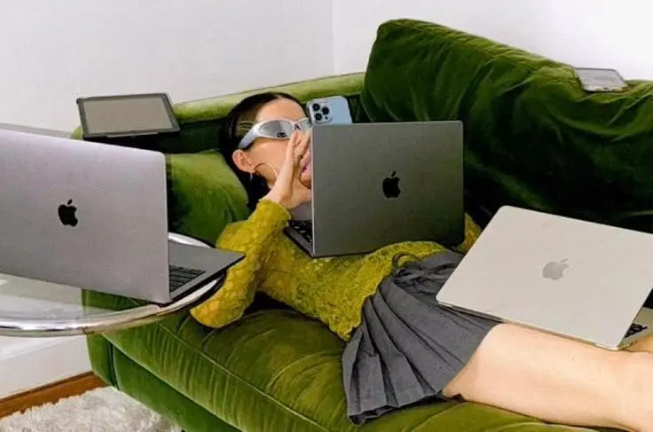 Girl in techwear on green couch surrounded by Apple laptops