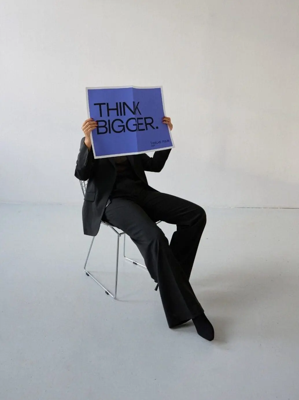 Person in a black suit holding a poster that says “THINK BIGGER” while sitting in a minimal white space