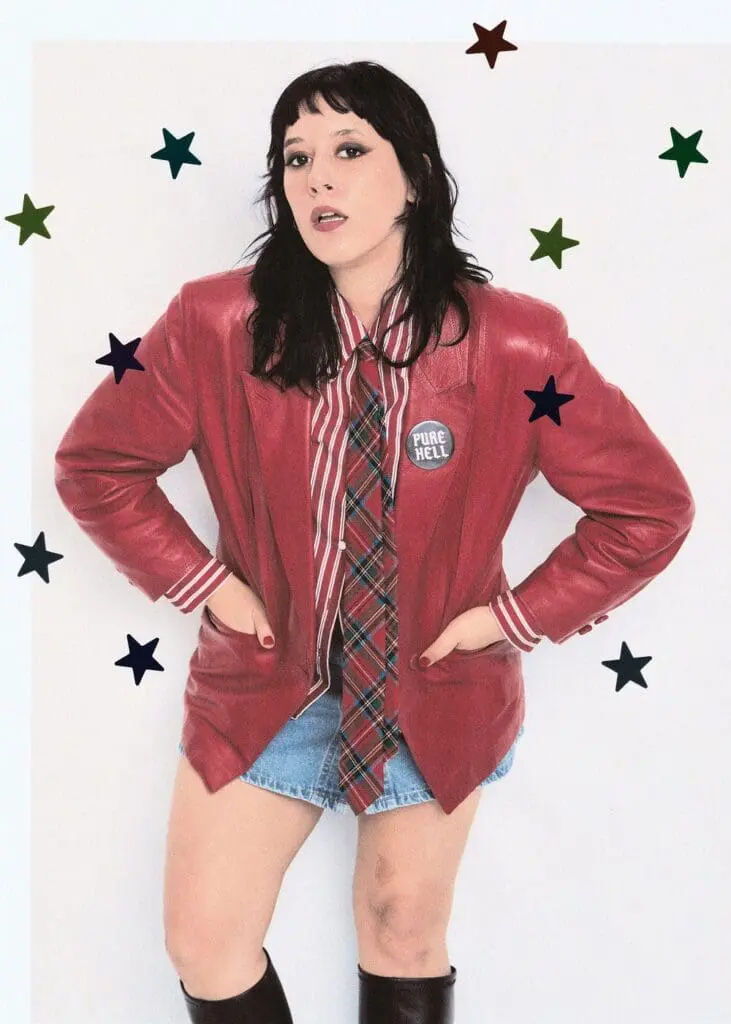 Güner Künier standing in red leather jacket, plaid tie, denim skirt and knee-high black boots, with decorative stars on background