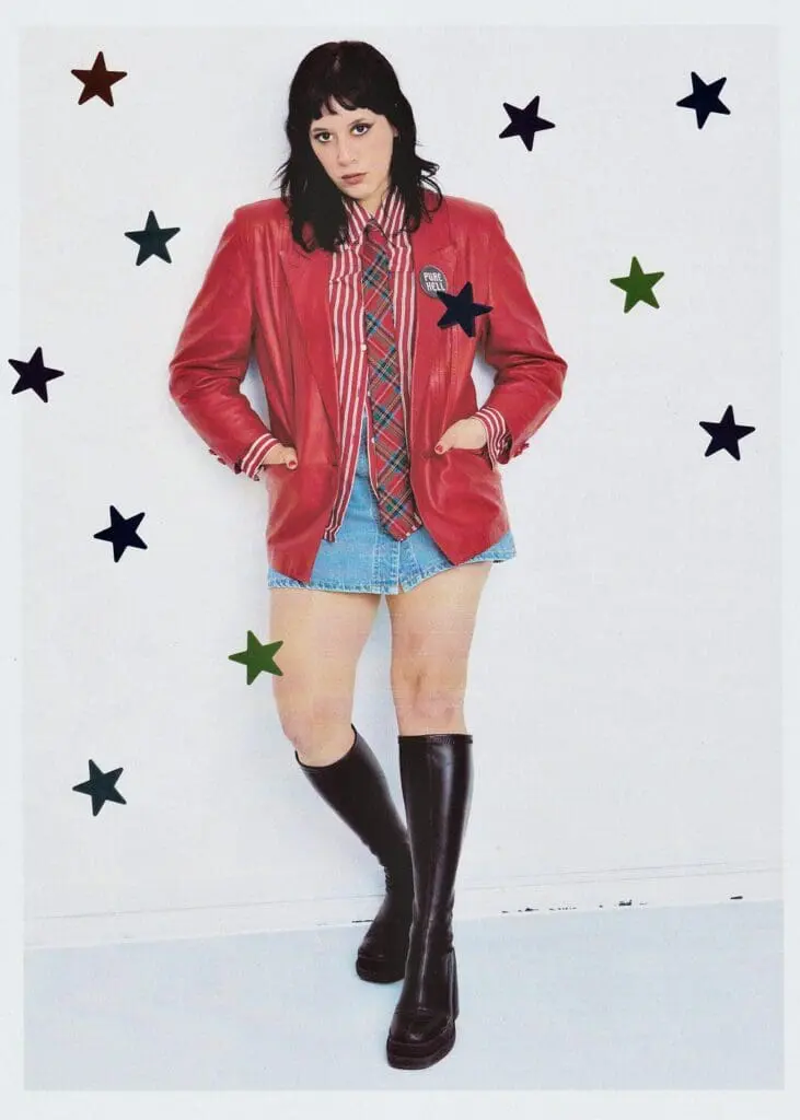 Güner Künier staring directly at camera in red leather jacket and tartan tie, with stars on the wall behind her