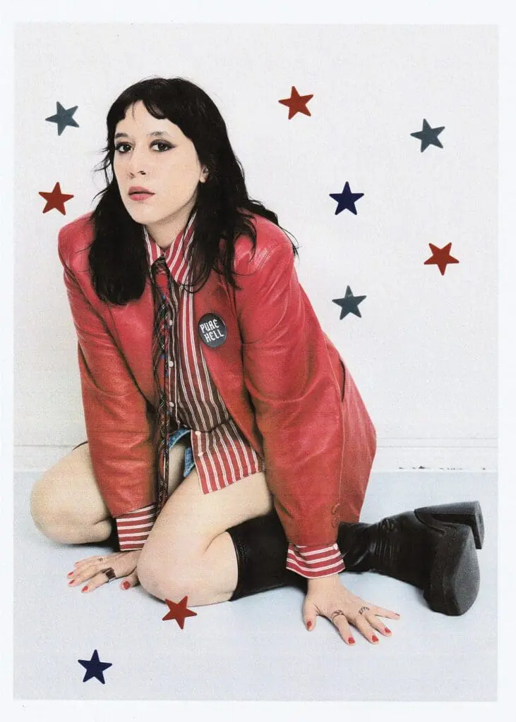 Güner Künier kneeling in a red leather jacket and striped shirt with star stickers on a white wall