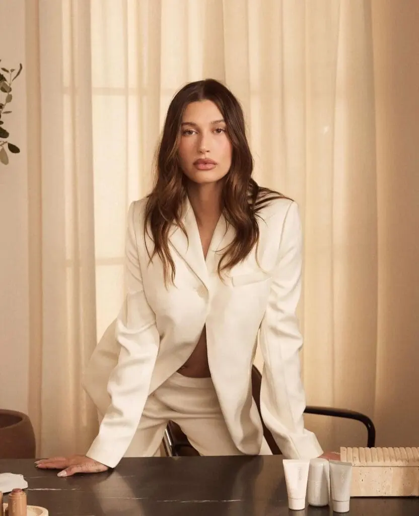 Hailey Bieber in a white suit standing behind a table of Rhode products. Brand identity