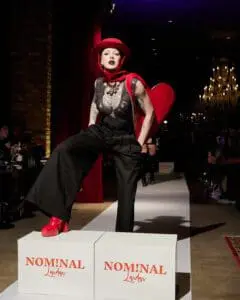Model in theatrical black jumpsuit, red heart hat and boots on runway