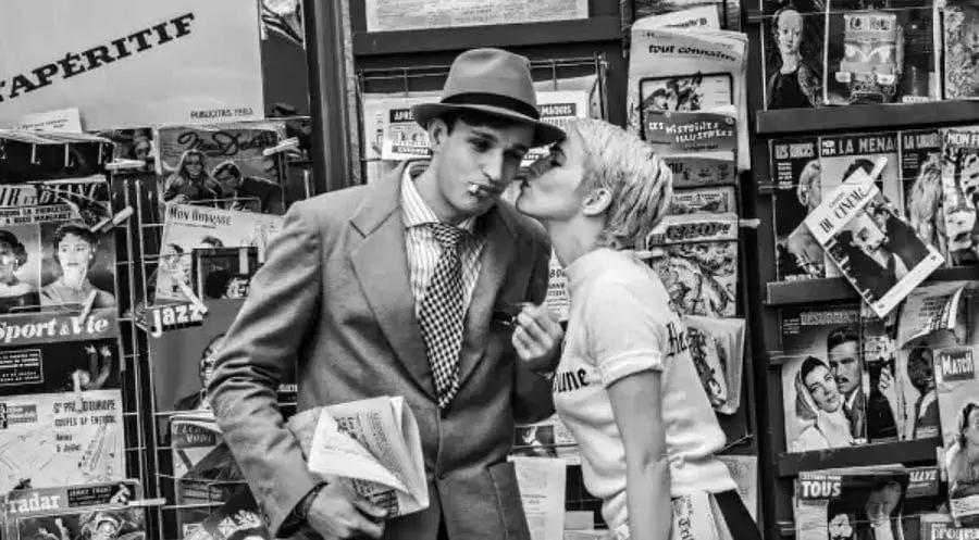 Young man in a suit being kissed on the cheek by a woman in front of a magazine kiosk