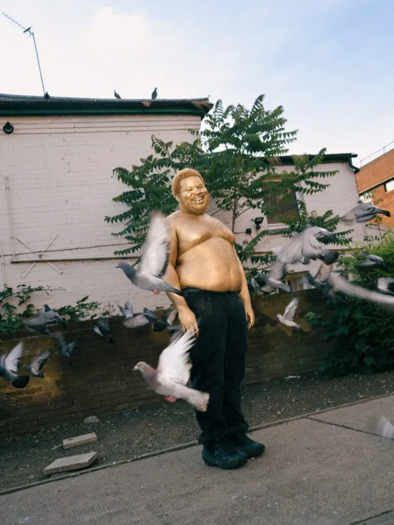 Shirtless golden-painted figure surrounded by flying pigeons