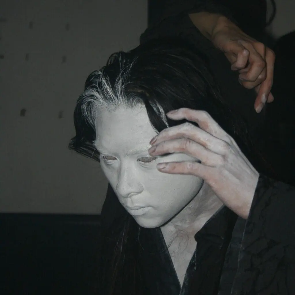 Performer with long hair and white-painted face mid-performance, adjusting hands