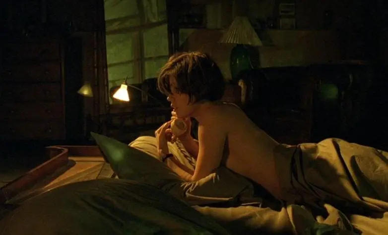 Young woman lying topless under sheets, speaking into a rotary phone in dimly lit room