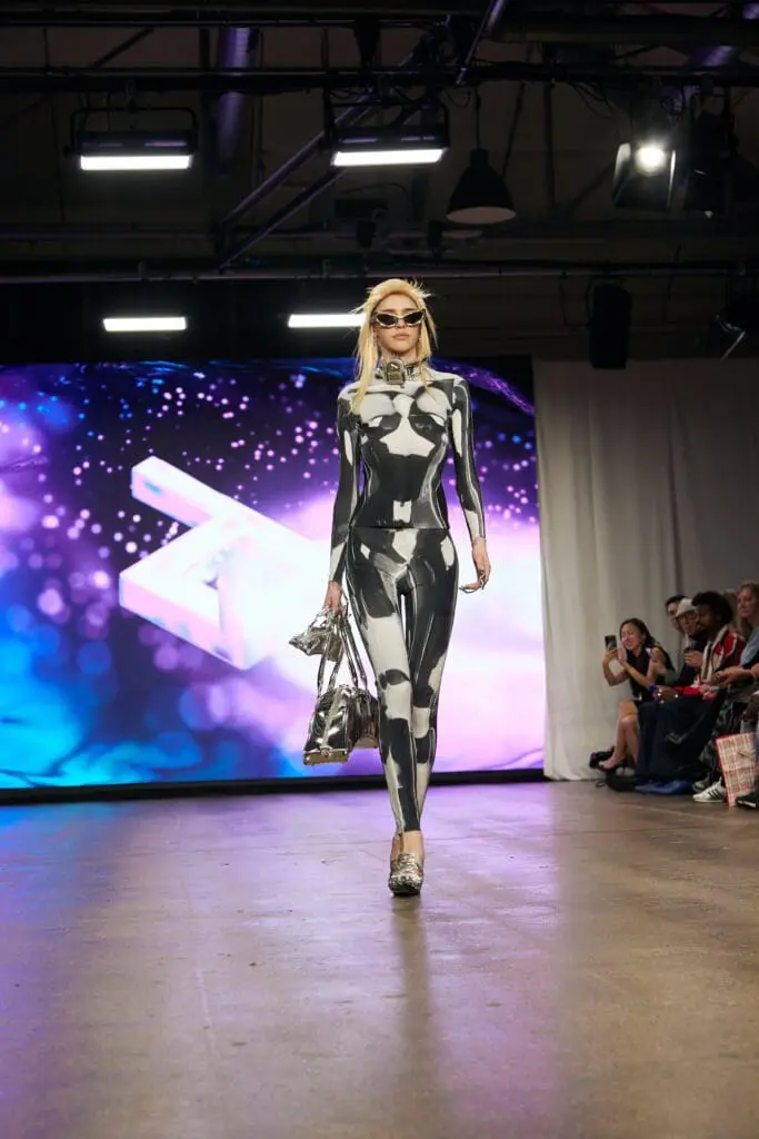 Model in a monochrome optical-print bodysuit carries metallic silver bags on the ZIDI runway at LFW.