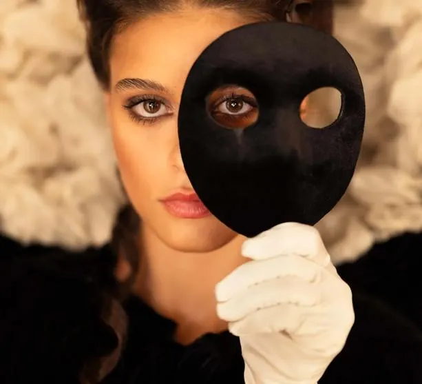 Close-up of a woman holding a black mask with two eye holes in front of one eye, highlighting performance of identity